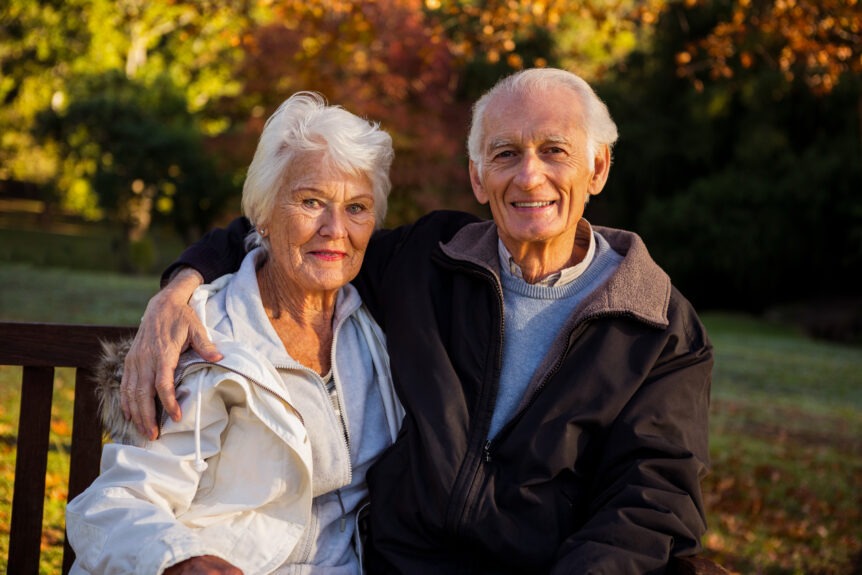 Happy retired couple sitting on a bench outdoors during autumn, smiling confidently as they enjoy their secure and peaceful retirement.